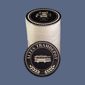 Es ist eine Packung mit 100 Bierdeckeln zu sehen. an die Packung angelehnt ist ein einzelner Bierdeckel auf dem man das Tramdepot Logo erkennen kann. Der Bierdeckel ist schwarz und das Logo ist hellbraun.