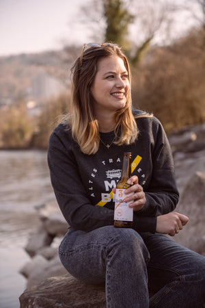 Frau mit Bier in der Hand sitzt mit Hellem Lagerbier in der Hand an der Aare. Sie trägt einen Pullover mit dem 