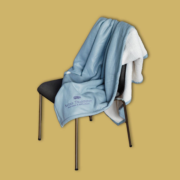 Decke über Stuhl gehängt. Hinten ist Decke weiss uns sehr flauschig. Vorne ist sie blau und ganz weich. Das Tramdepot Logo ist auf der Vorderseite draufgestickt.